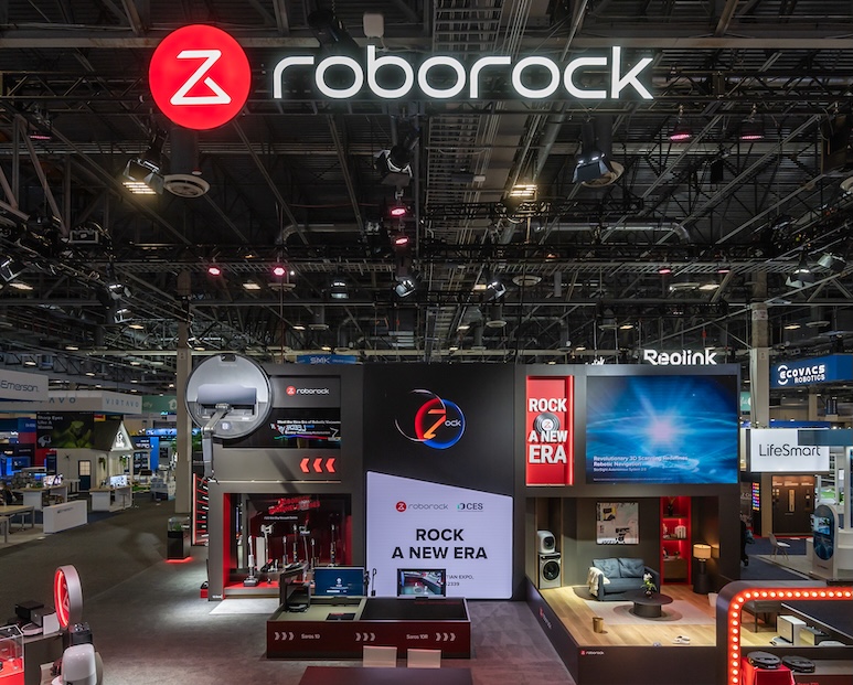 Front view of the Roborock booth at CES 2025, featuring a large illuminated brand logo overhead and a central digital screen displaying the theme “Rock a New Era.” The booth design includes a suspended robotic vacuum model, a lifestyle living room setup, and various product interaction zones. Bright red accent lighting and bold typography emphasize Roborock’s futuristic and design-driven identity.